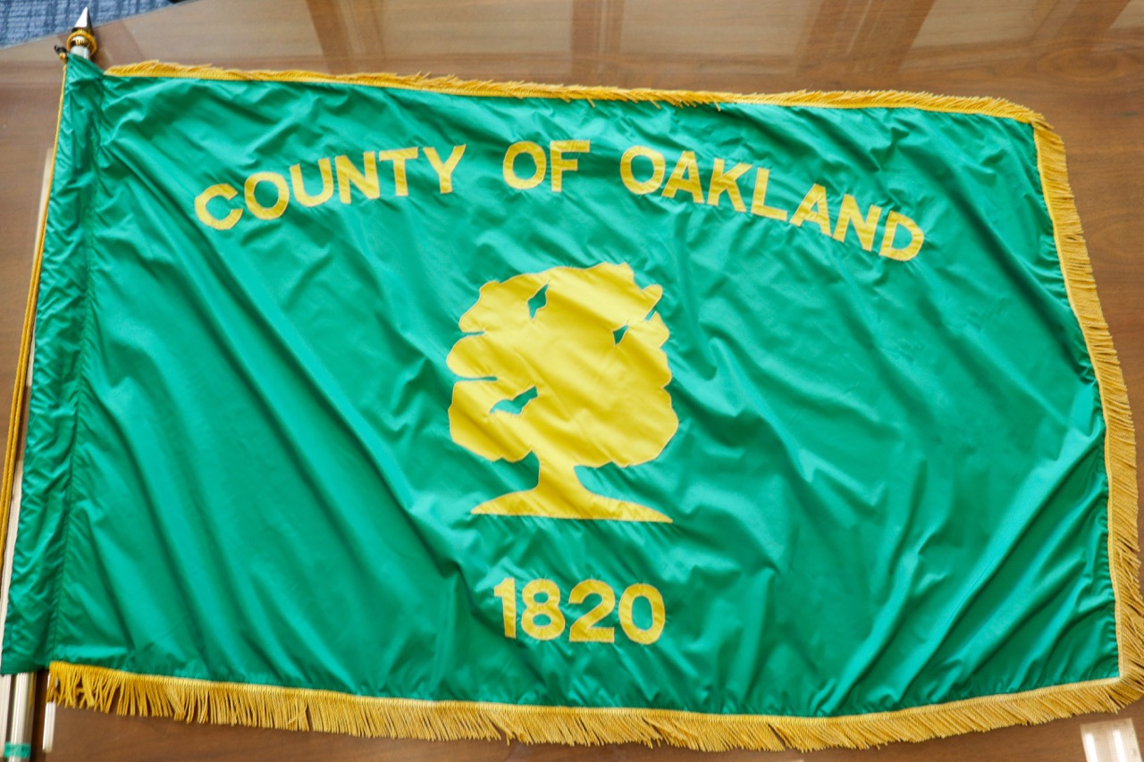 Photo of the current Oakland County Flag. A green flag with yellow text that says
