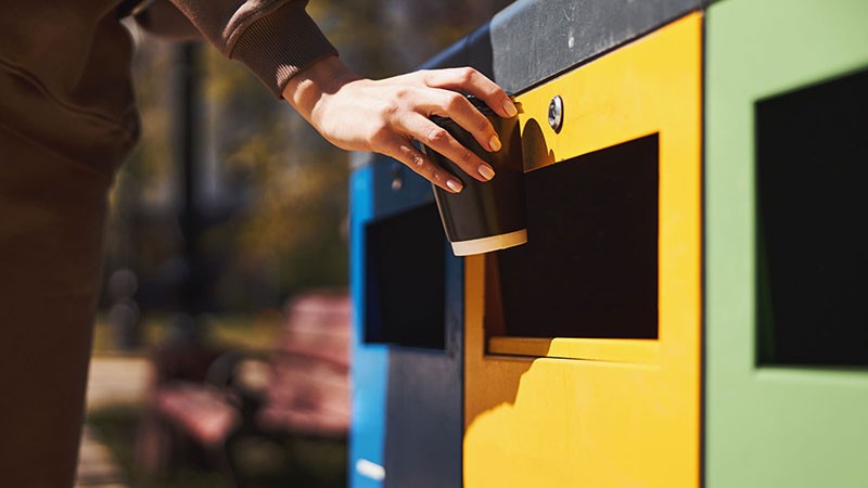 A person placing a paper cup into a yellow recycling bin, with blue and green bins visible beside it. The scene is outdoors with a bench and blurry background.