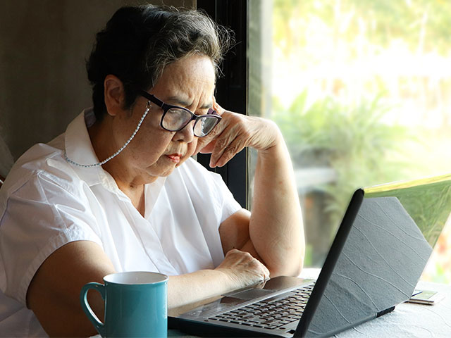 Asian senior woman sitting by the window with blue cup of coffee and computer laptop on table, looking at computer screen.