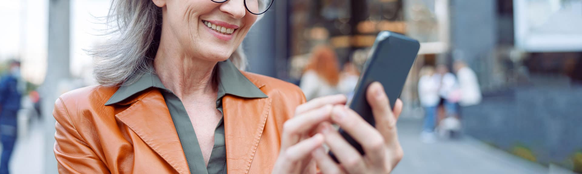 Positive mature lady with glasses uses mobile phone standing on modern city street on autumn day.