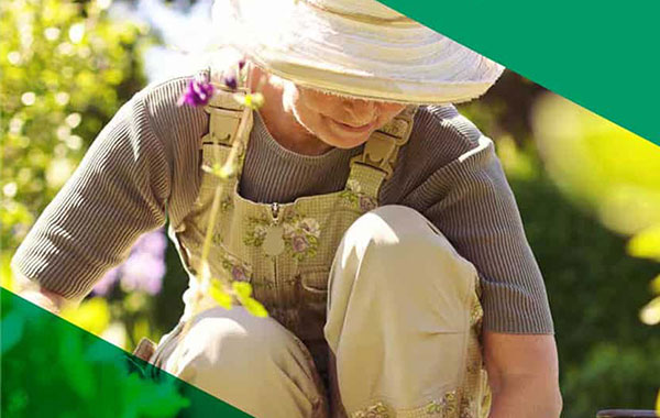 Older looking woman working in a garden with flowers around her wearing a hat.