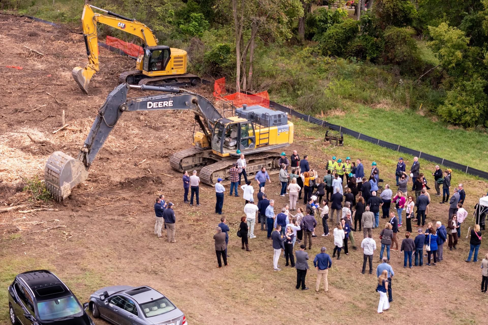 A group of people gathers around two large excavators on a cleared construction site, bordered by green trees. The mood is sociable and focused.