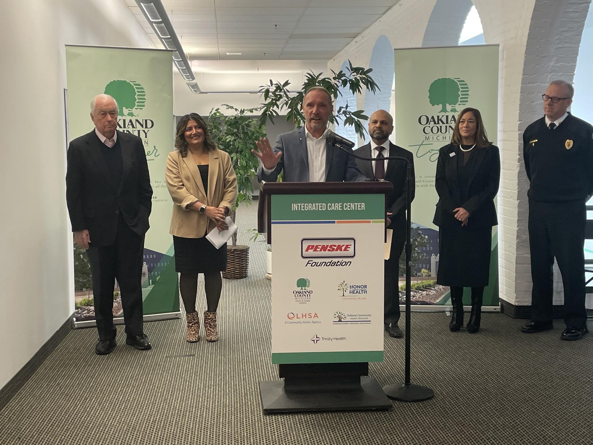 Several people gather at a podium, speaking to an audience with a green sign displayed behind them.