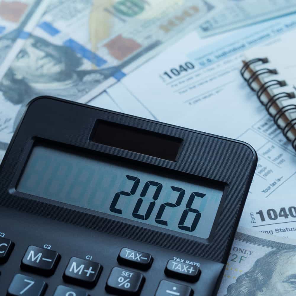 A calculator displays 2026 atop U.S. tax forms and a spiral notebook, surrounded by hundred-dollar bills, conveying financial planning themes.