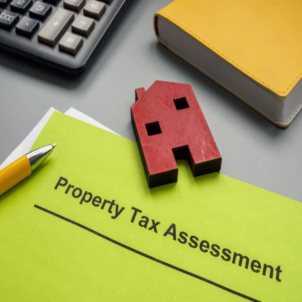 Yellow document labeled 'Property Tax Assessment' on a desk with a pen, calculator, book, and red house model, suggesting a financial review context.